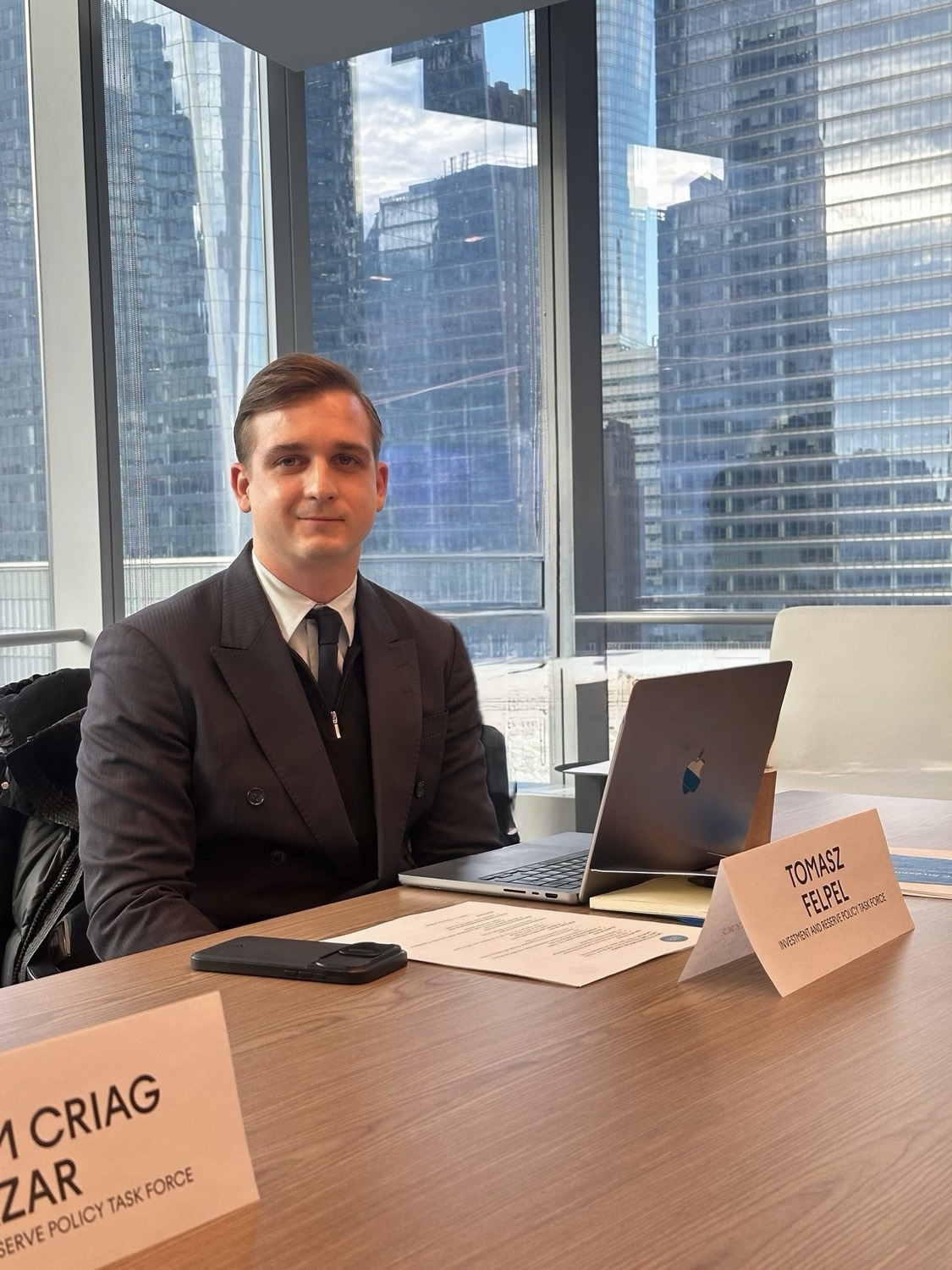 Tomasz Felpel seated at a boardroom table with a Reserve Policy Task Force name placard, the New York skyline behind him.